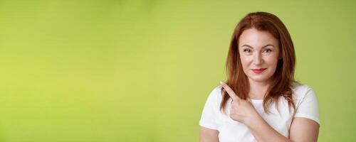 Kind cheerful good-looking middle-aged woman 50s redhead white t-shirt smiling modest assured give advice pointing upper left corner indicating great promotion advertising product green background photo