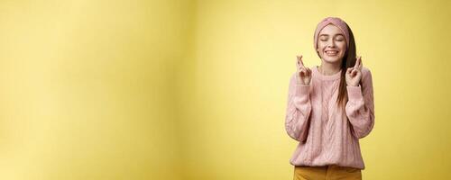 Girl wishing wellness wanting dream come true, cross fingers dreamy, close eyes waiting miracle, anticipating good news, posing excited and joyful against yellow background in knitted warm sweater photo