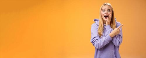 Amazed excited overwhelmed young blond girl in hoodie press palm cheek screaming astonished pointing right seeing famous super star cannot hold joyful positive emotions, orange background photo