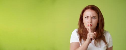 Not speak during exam. Strict serious-looking displeased middle-aged redhead woman frowning disappointed hushing say shush index finger pressed lips keep quiet gesture green background photo