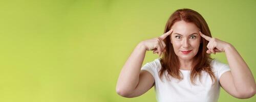 Woman playfully staring funny control your mind. Silly redhead middle-aged female touch temples popping eyes smiling delighted read thoughts trying guess intrigued what thinking green background photo