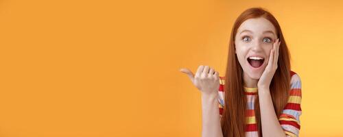 Cheerful excited impressed talkative girl pointing astonished thumb left drop jaw smiling broadly touch cheek thrilled, interesting event discuss awesome wonderful performance, orange background photo