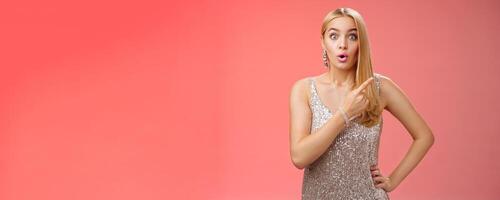 Wondered amused attractive blond woman talking party widen eyes say wow pointing questioned right direction see famous person standing surprised impressed gaze camera, red background photo