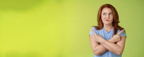 Time make right choice. Perplexed hesitant redhead middle-aged female customer deciding between few variants cross arms body pointing sideways look left smirking unsure doubting choosing product photo
