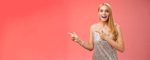 Excited carefree happy young attractive woman joyful attend awesome party in silver beautiful glittering dress pointing left suggesting come together inviting dance smiling cute red background photo