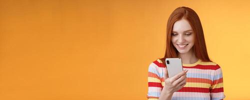 Charming modern redhead girl college student checking message box holding smartphone look happy smiling delighted cellphone display receive hundred likes photo post online, orange background