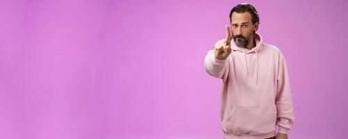One rule listen. Serious-looking bossy focused determined adult bearded male in pink hoodie extend index finger first number scolding making statement forbid bad behaviour, purple background photo