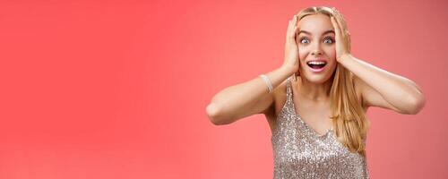 Amused surprised excited young blond attractive woman in silver glittering stylish dress hold head hands widen eyes speechless look camera unbelievable thrill fan see famous person, red background photo