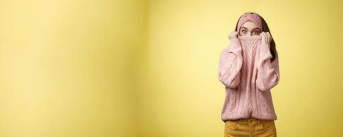 Girl hiding in collar wearing knitted sweater over nose popping eyes, staring bugged eyes at camera, hiding face reacting amazed and shocked, feeling scared, standing in stupor over yellow background photo