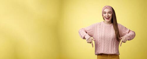 Euphoric attractive young trendy girl in knitted band wearing sweater grinning excited enthusiastic pointing down thrilled of awesome promotion standing amused and overwhelmed over yellow background photo