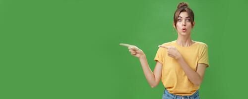 Woman reacting to awesome sales in near shop pointing left and gazing impressed and excited at camera folding lips from desire and amazement staring at camera thrilled posing over green background photo