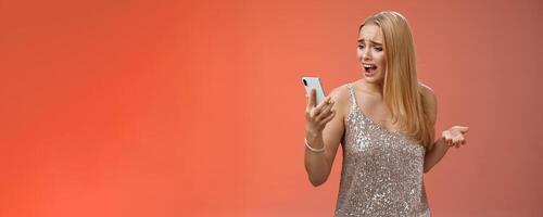 Troubled concerned arrogant young blond woman complaining yelling smartphone cannot call friend no signal holding smartphone look mobile display pissed moody arguging, red background photo