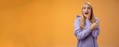 Amazed excited overwhelmed young blond girl in hoodie press palm cheek screaming astonished pointing right seeing famous super star cannot hold joyful positive emotions, orange background photo