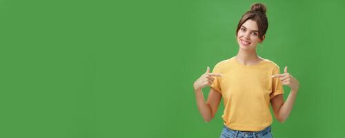Calm and polite friendly-looking skillful young female coworker in yellow t-shirt pointing at herself self-assured tilting head and smiling at camera wanting participate in event over green background photo