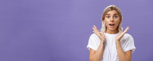 Close-up shot of charmed and impressed good-looking caucasian girlfriend with blond hair and tattoos on arms raising hands in amazed and excited gesture opening mouth and gazing mesmerized at camera photo