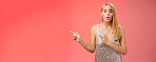 Intrigued excited attractive tender feminine blond european girl in silver stylish dress pointing left amused talking camera widen eyes thrilled asking boyfriend wanna dance shall go check out promo photo
