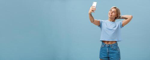New post in my blog. Stylish feminine and sociable good-looking young female posing for selfie with pulled arm and smartphone sticking out tongue while making faces at device screen over blue wall photo