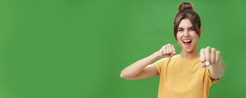 Cute female rebel in yellow t-shirt with gap teeth pulling fist towards camera as if showing fighting skills yelling daring and excited standing over green background smiling acting like boxer photo