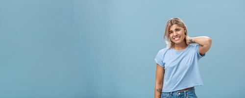 Girl unwilling to go with friend feeling awkward and unsure how say no frowning and clenching teeth making apologizing face rubbing neck behind saying sorry while rejecting offer over blue background photo