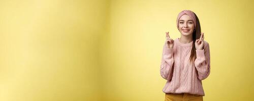 Cross my fingers for you. Supportive cute young european girlfriend wearing sweater, glamour headband making luck, wish gesture smiling hopeful having faith dream come true over yellow background photo