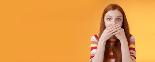 Oops sorry. Girl feeling awkward saying inappropriate word standing unconfident cover mouth palms wide eyes staring camera shocked express surprise nervously standing orange background photo