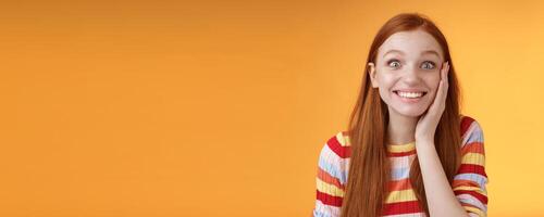 Cheerful glad excited attractive redhead girl blushing surprised feeling happy touch cheek pleased receive good news standing joyful thrilled get awesome chance, posing orange background photo
