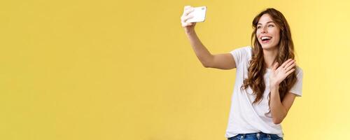 Friendly lively good-looking cheerful feminine girl extend arm hold smartphone record blog waving front camera smiling broadly talking fans videocalling taking selfie yellow background photo