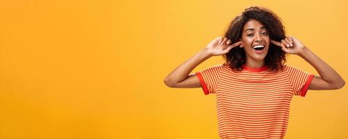 Do not care what saying cannot hear you. Carefree indifferent good-looking african american woman with curly hairstyle closing ears with index finger singing lalala during argument behaving childish photo