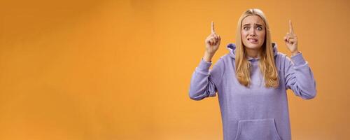 Worried perplexed unsure cute blond woman presenting project standing awkward afraid humiliation crining scared pointing up raised index finger waiting reply nervously, orange background photo