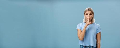 Promise not tell anyone it secret. Mysterious attractive and feminine blond woman with tanned skin and tattoos shushing with index finger over mouth squinting as if hiding something over blue wall photo