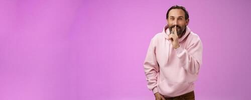 Lips sealed secret. Portrait alluring cheeky stylish adult bearded guy in pink hoodie asking keep quiet show shush shh gesture index finger pressed mouth smiling devious smug face, have idea photo