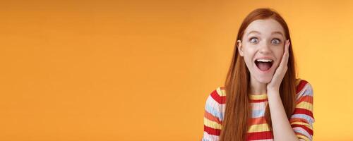 Amused happy smiling good-looking redhead woman screaming happily touch cheek astonished receive good perfect news triumphing feeling excited look thrilled camera, standing orange background photo