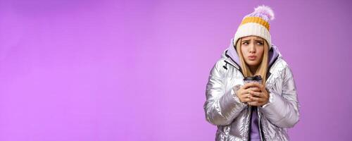 Trembling uncomfortable cute pouting young blond girl feel freezing cold winter snowy weather outside shaking low temperature hold take-away hot coffee cup warming up, purple background photo