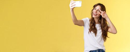 Joyful charismatic good-looking curly-haired woman white t-shirt show peace victory sign smiling broadly posing tender friendly expression smartphone front camera take selfie yellow background photo