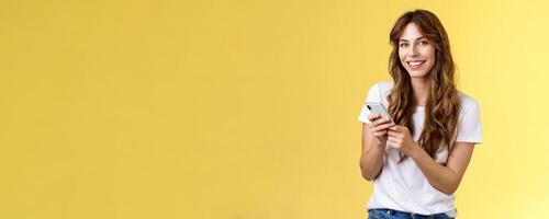 Enthusiastic good-looking urban girl wear white t-shirt standing casual smiling delighted camera texting hold smartphone scroll social media feed stand yellow background browsing network photo