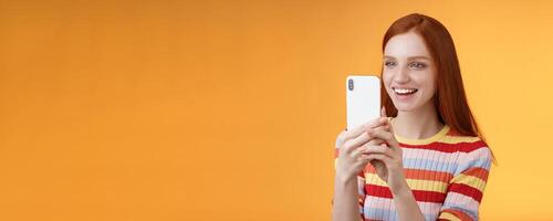 Redhead girl having fun recording hilarious friend actions hold smartphone look display amused shooting funny telephone standing orange background satisfied smiling delighted photo