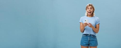 So happy receive premium for free reacting with enthusiasm and excitement on amazing message smiling broadly with delight and surprise holding smartphone posing over blue background photo