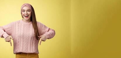 Euphoric attractive young trendy girl in knitted band wearing sweater grinning excited enthusiastic pointing down thrilled of awesome promotion standing amused and overwhelmed over yellow background photo