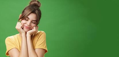 Tender sensual timid european girl in yellow t-shirt leaning head on shoulder touching cheeks with hands closing eyes and smiling with soft grin feeling nostalgic and romantic over green background photo