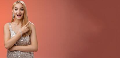 Joyful surprised good-looking blond woman talking friend during party smiling broadly look camera amused wondered pointing upper right corner curious discuss awesome room design, red background photo