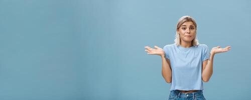No idea how help sorry. Portrait of unsure gloomy girlfriend feeling regret shrugging with hands near shoulders frowning and pursing lips being uncertain and unaware over blue background photo