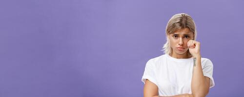Studio shot of offended sad and timid silly woman with blond hairstyle frowning looking from under forehead holding fist near eye as if whiping teardrop being upset over purple background photo