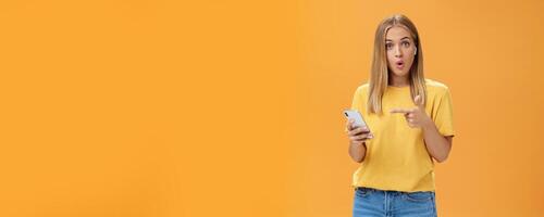 Wman impressed by wireless earphones connecting to smartphone themselves pointing at device screen questioned and surprised folding lips looking intrigued at camera wearing earbuds over orange wall photo