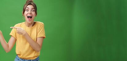 Waist-up shot of enthusiastic and charismatic impressed happy girl laughing and smiling broadly pointing left at funny hilarious thing reacting surprised and entertained, posing over green background photo