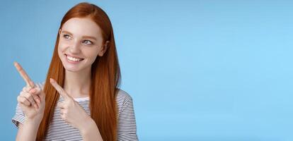 Attractive redhead female model pure clean skin ginger long hair pointing looking left intrigued smiling satisfied indicating interesting advertisement intrigued using new product, blue background photo