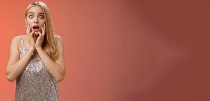 Shocked concerned stunned young european blond woman in silver dres drop jaw widen eyes stunned speechless gasping open mouth touch face amazed, look troubled perplexed, red background photo