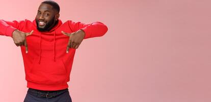 Sassy young charismatic handsome bearded black guy in red hoodie pointing down index fingers smiling broadly showing awesome copy space solution cool product for you, standing pink background photo