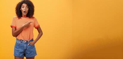 Studio shot of stylish fit female model in denim shorts and t-shirt gasping impressed pointing right while asking question being interested during observing masterclass over orange background photo