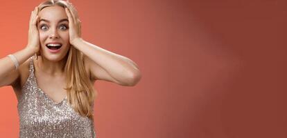 Amused surprised excited young blond attractive woman in silver glittering stylish dress hold head hands widen eyes speechless look camera unbelievable thrill fan see famous person, red background photo