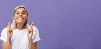 Waist-up shot of hopeful optimistic attractive stylish woman in white t-shirt with tattooed arms winking smiling joyfully while crossing fingers for good luck making wish over purple background photo